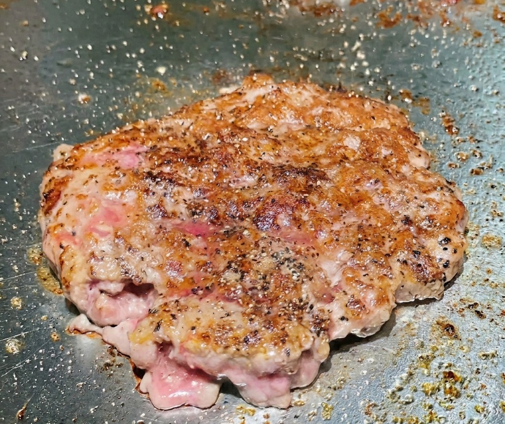 A flipped beef patty on a grill showing a deeply seared and browned crispy surface.