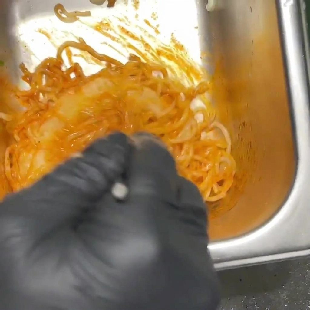 Spicy orange ramen noodles being mixed in a metal container by a person wearing black gloves.