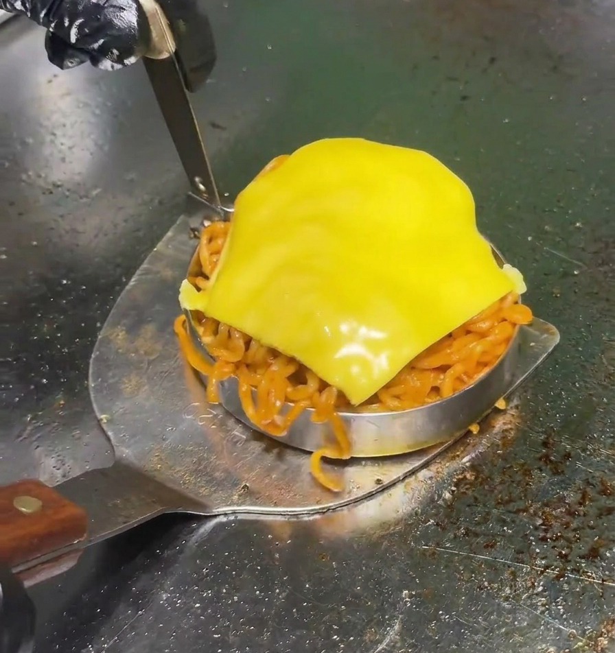 A metal spatula lifting a circular mold filled with spicy red noodles and topped with a slice of yellow cheese on a professional grill.