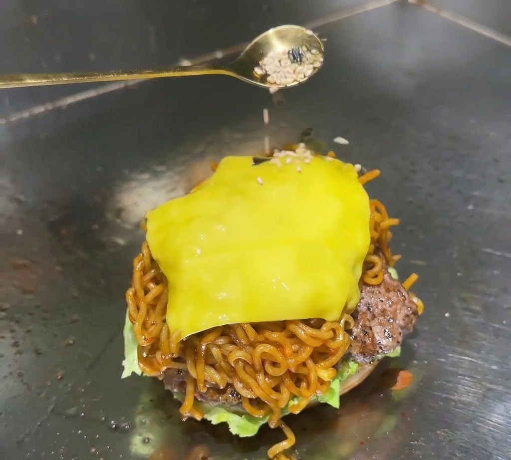 A golden spoon sprinkling a mixture of white sesame seeds and black seaweed flakes onto the melted cheese topping of a ramen burger.