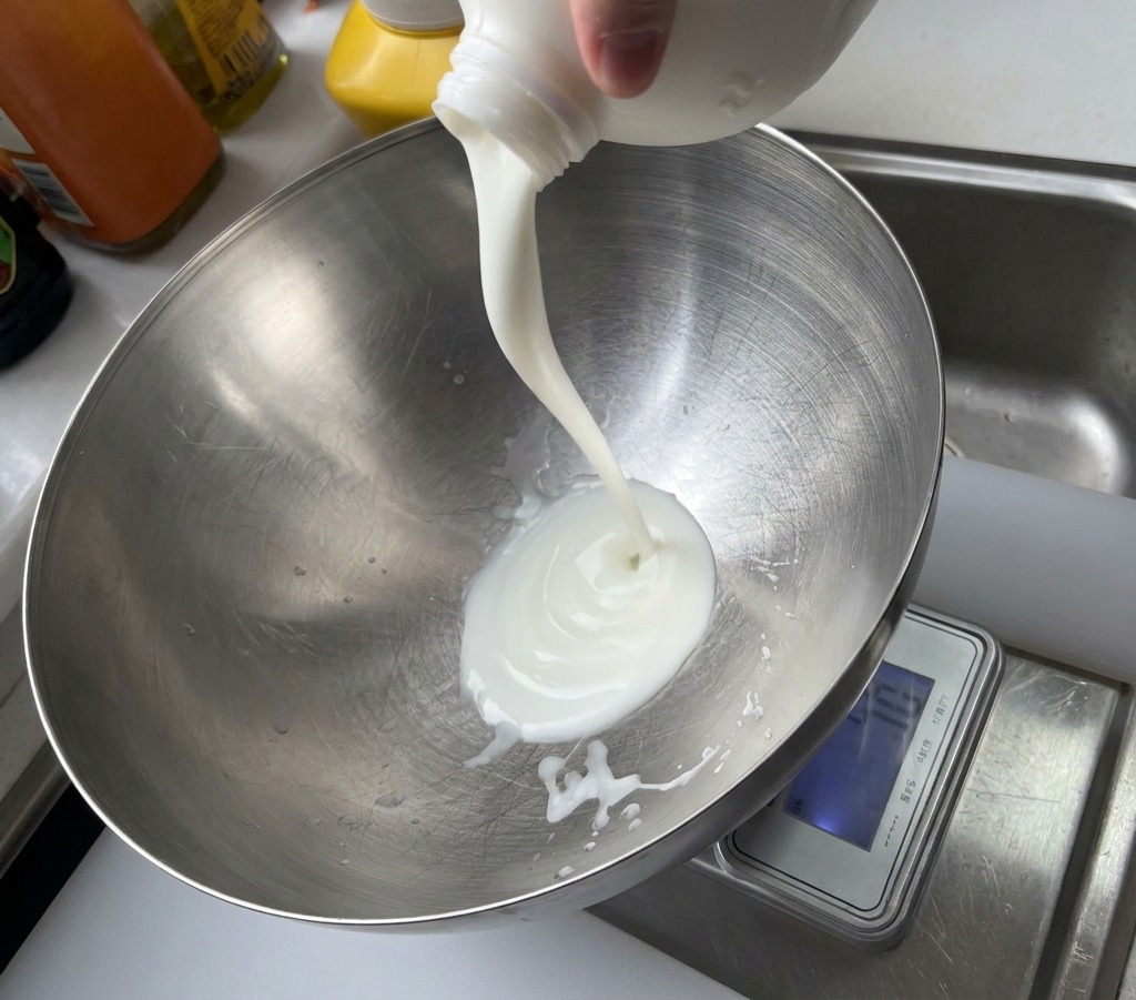 A stream of white yogurt being poured from a plastic container into a large, brushed metal mixing bowl.