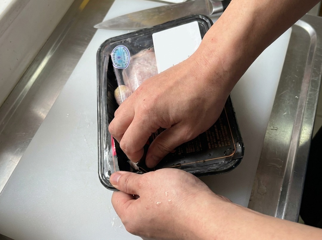 Hands opening a plastic-wrapped tray of raw chicken thighs on a white cutting board next to a kitchen knife.