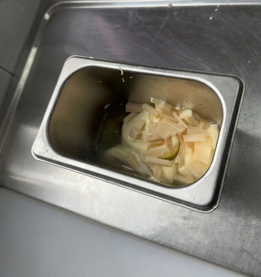 A stainless steel container holding a scoop of mayonnaise, yellow mustard, and small bits of chopped green pickles.