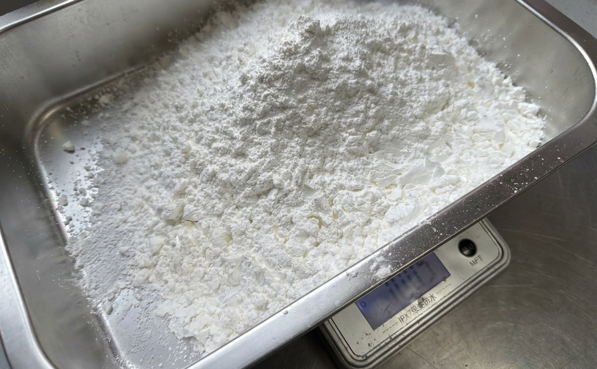 A large pile of white cornstarch being weighed on a digital scale inside a rectangular stainless steel basin.