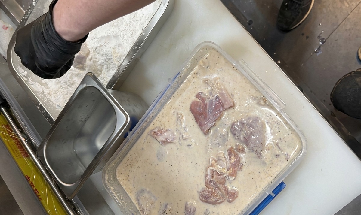 Gloved hands transferring chicken marinated in a milky liquid into a tray of seasoned breading flour.