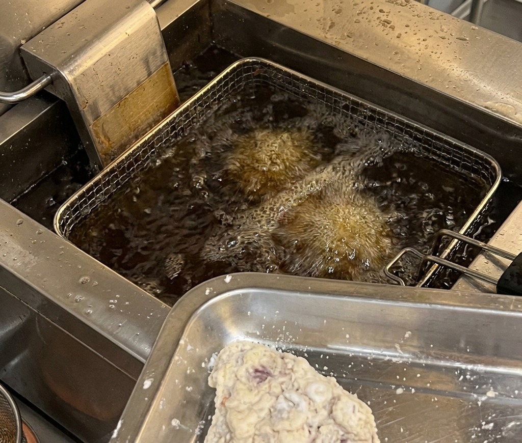 Breaded chicken pieces on a metal tray being prepared to enter a commercial deep fryer with hot, bubbling oil.