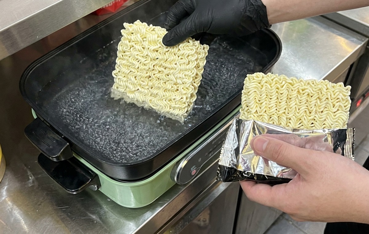 A person wearing black gloves dropping square blocks of dried instant noodles into a pot of boiling water.