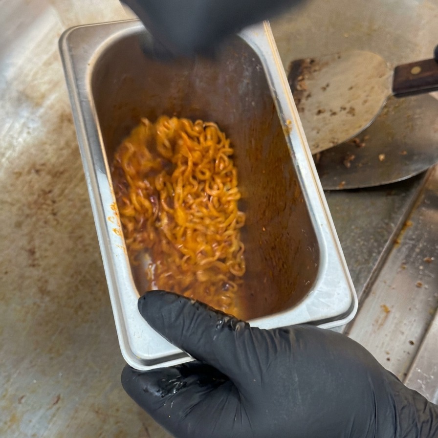 Spicy red noodles being mixed and tossed inside a deep stainless steel kitchen container.