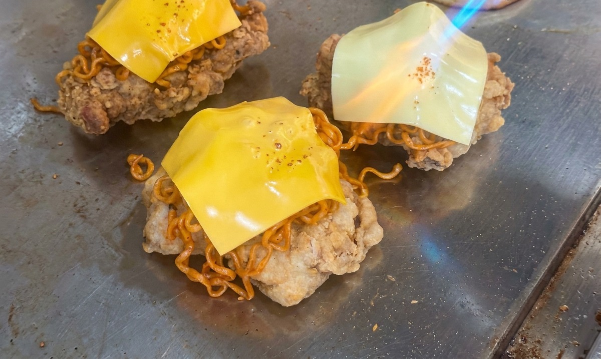 A blowtorch flame melting a square of yellow cheese over a pile of spicy noodles and fried chicken on a grill.