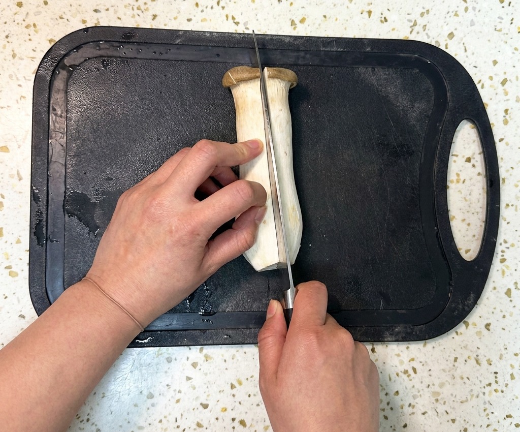 A person using a chefs knife to slice a large king oyster mushroom in half lengthwise on a black plastic cutting board.