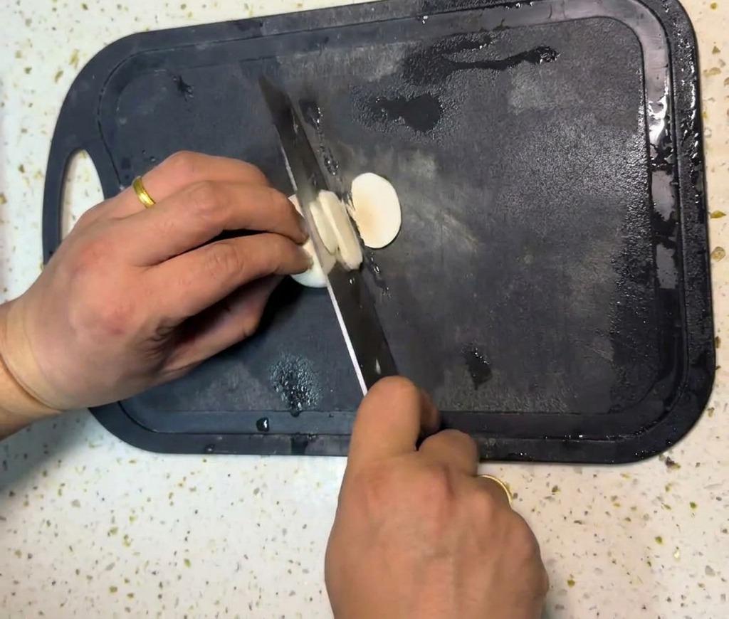 A persons hands using a chefs knife to slice white button mushrooms into thin rounds on a black cutting board.