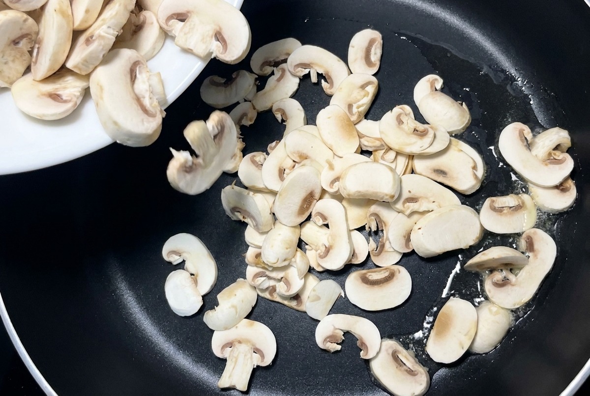 Sliced white mushrooms being poured from a white ceramic plate into a black non stick skillet.