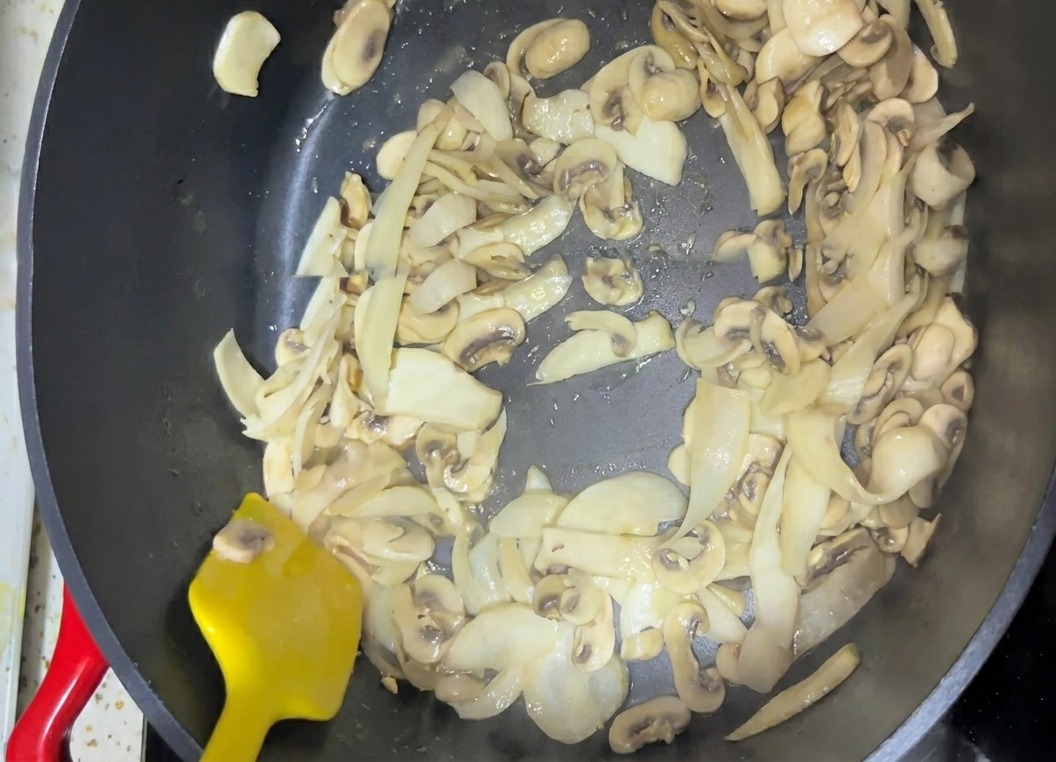 Sliced mushrooms being stirred in a dark cooking pan with a bright yellow spatula.