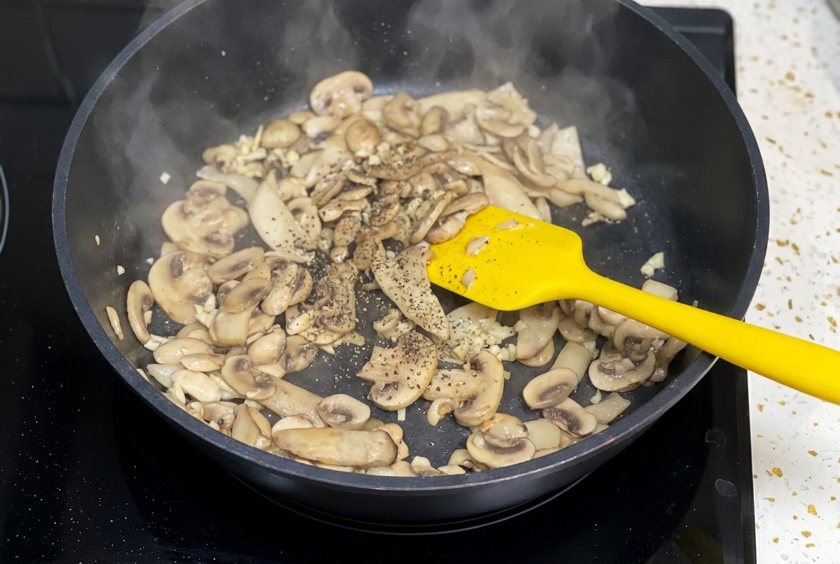 Sautéed mushrooms in a black skillet being seasoned with a dusting of ground black pepper.