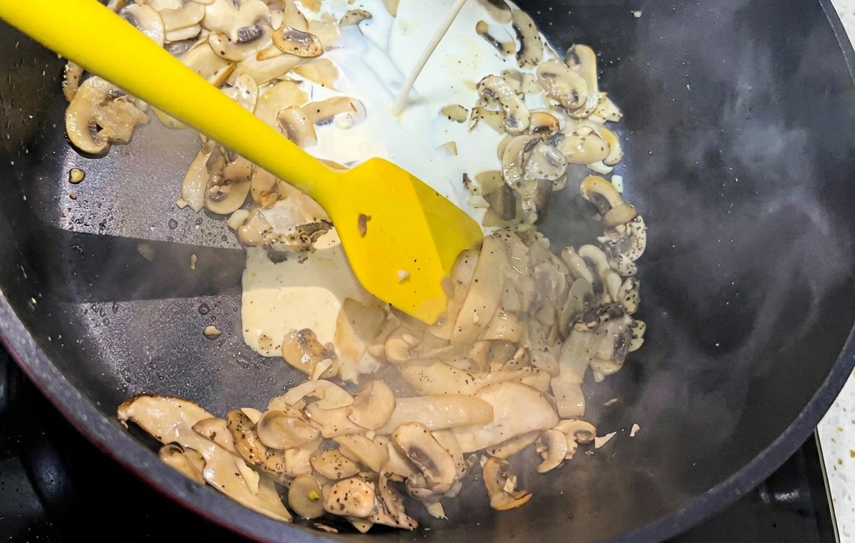 A stream of white heavy cream being poured into a black pan filled with sautéed mushroom slices.