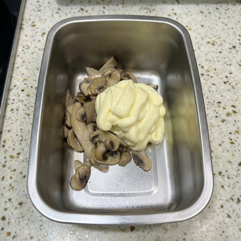 A metal container holding sautéed mushrooms topped with a large swirl of pale yellow mayonnaise.