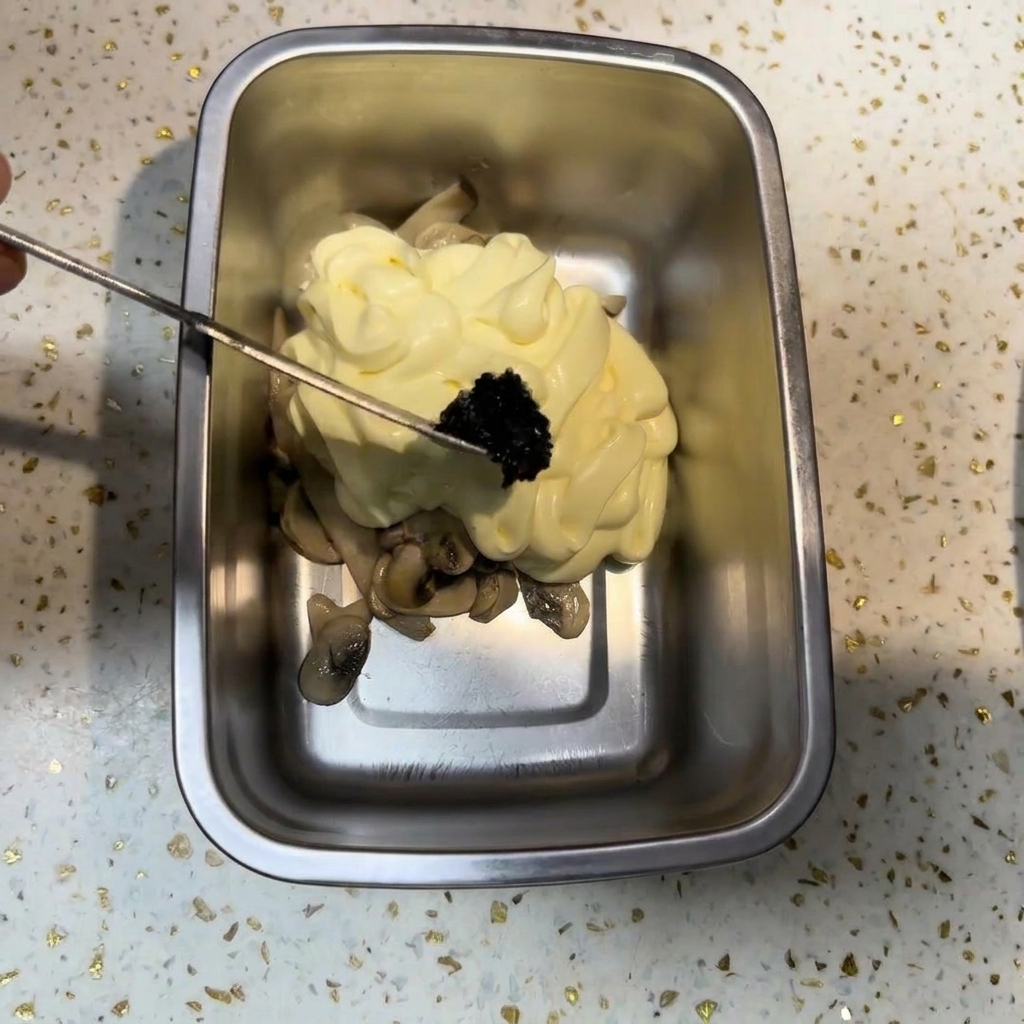 A small metal tool placing a portion of dark black truffle paste onto a mound of mayonnaise and mushrooms.