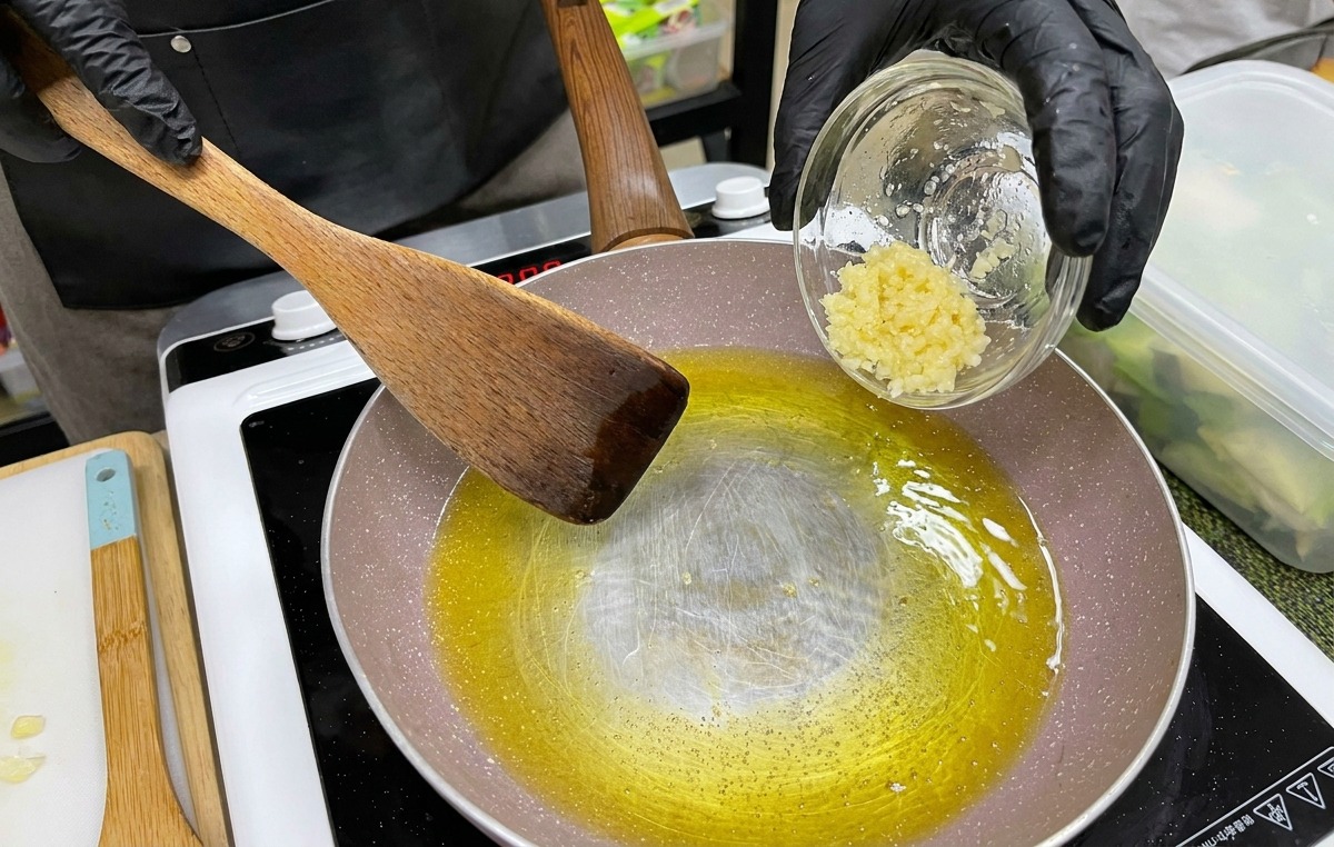 A hand in a black glove pouring a small bowl of minced garlic into a pan of melted yellow butter and oil.