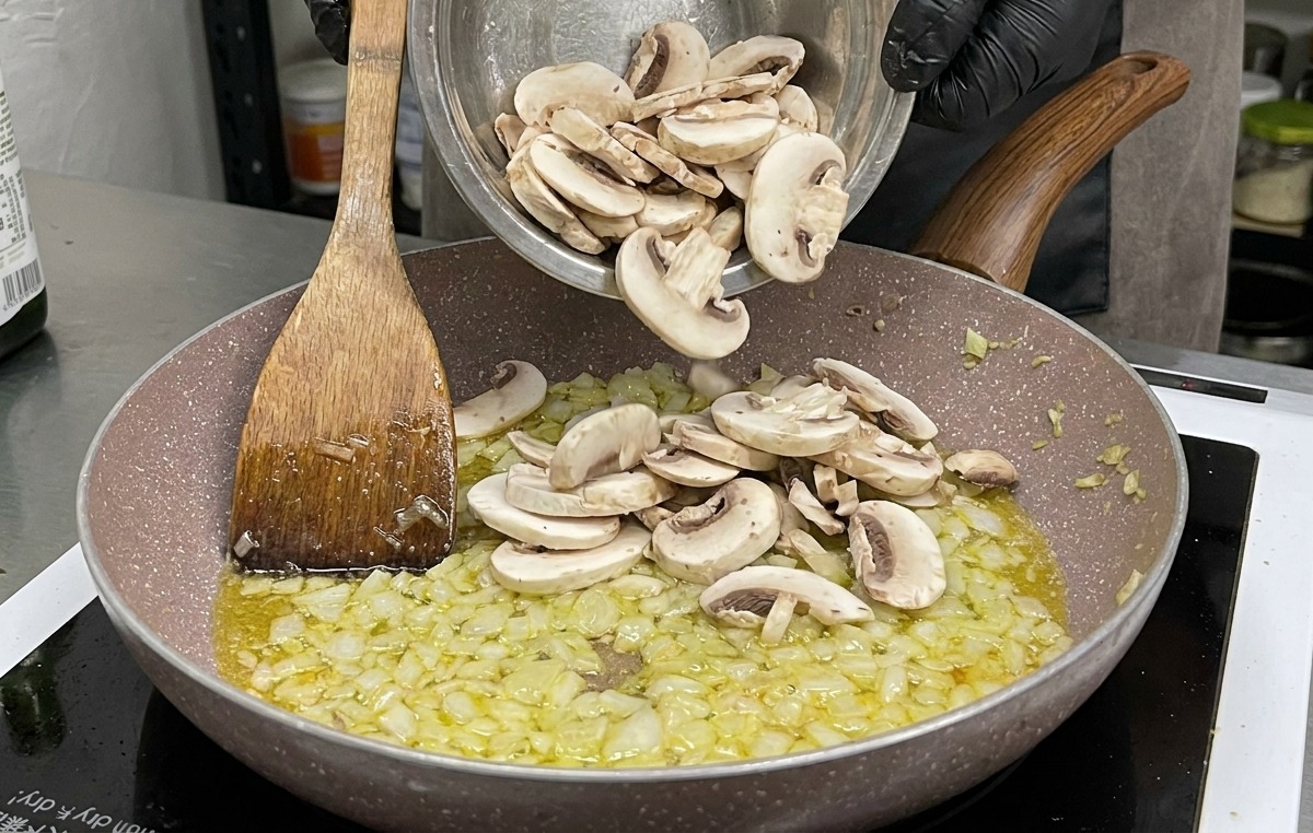 A large bowl of sliced raw white button mushrooms being added to the pan of sautéed onions and garlic.