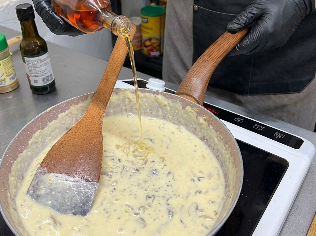 A person in black gloves pouring amber liquid from a glass bottle into a frying pan filled with creamy mushroom sauce.