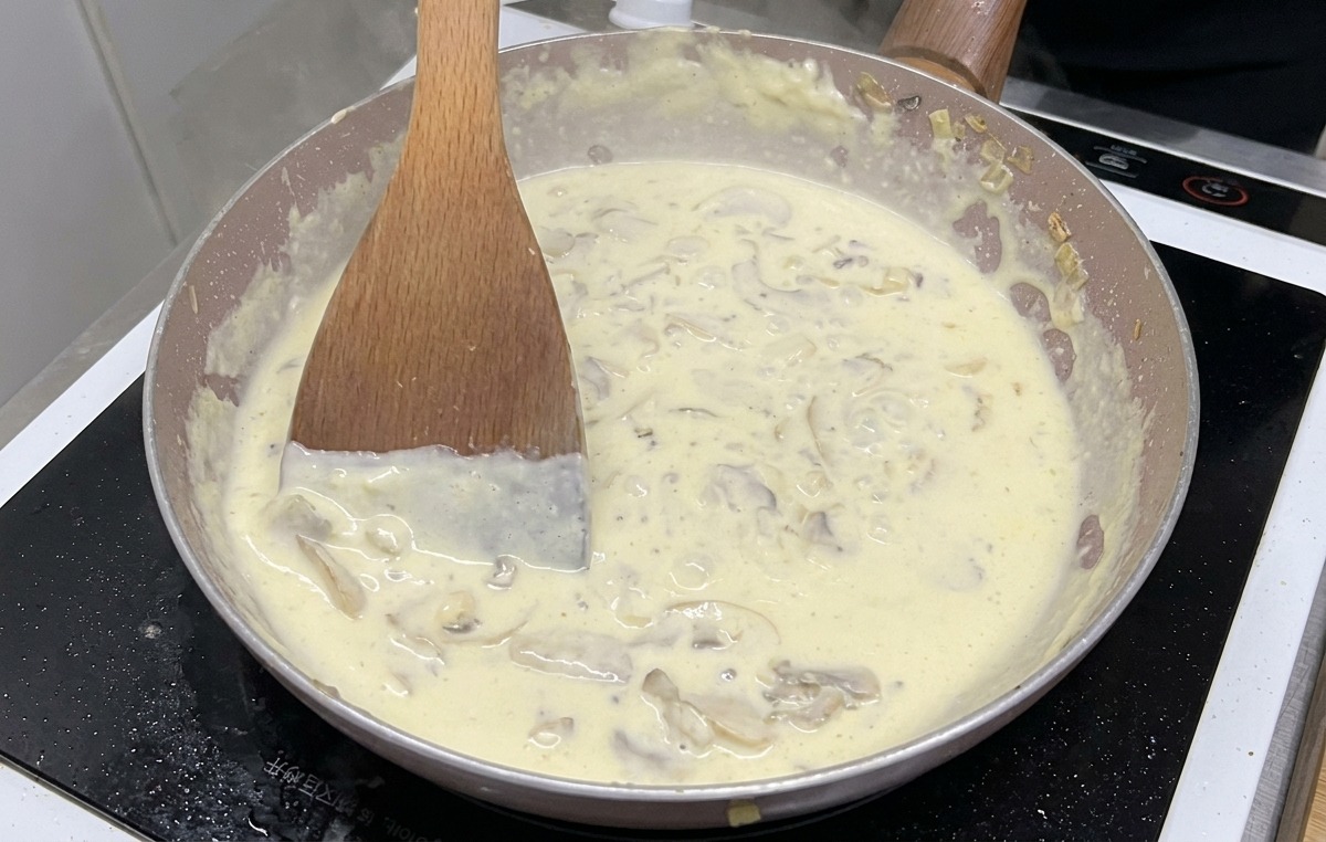 A creamy mushroom sauce with visible mushroom slices bubbling and simmering in a pan while being stirred with a wooden spatula.