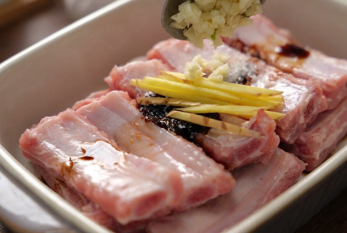 A spoon adding minced garlic onto seasoned pork ribs that are already topped with thin ginger matchsticks.