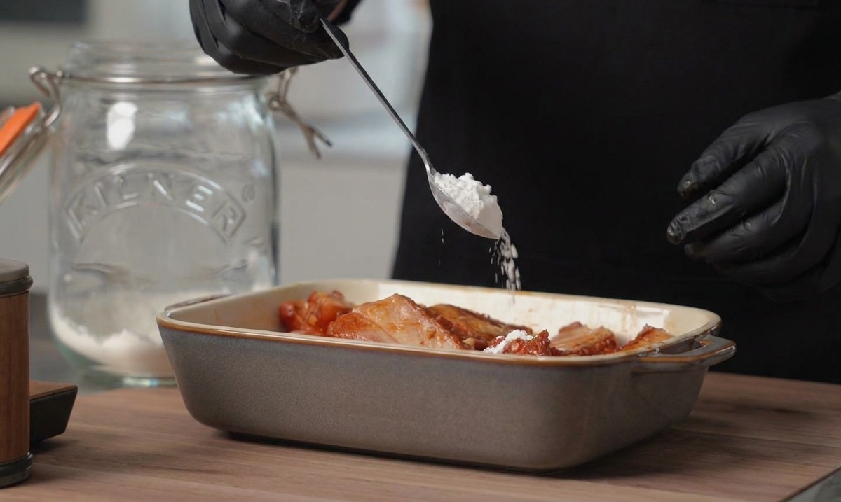 A person wearing black gloves adds a spoonful of white cornstarch to a rectangular dish filled with marinated pork ribs.