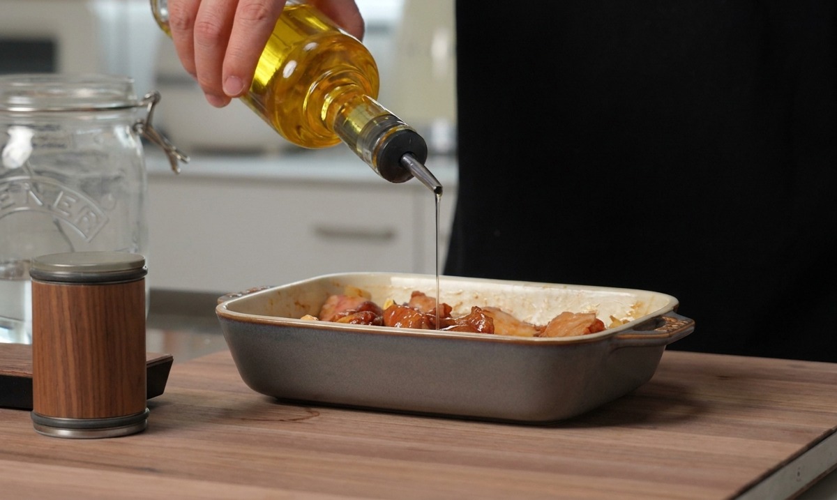 Golden cooking oil being poured from a glass bottle onto seasoned pork ribs sitting in a gray ceramic baking dish.