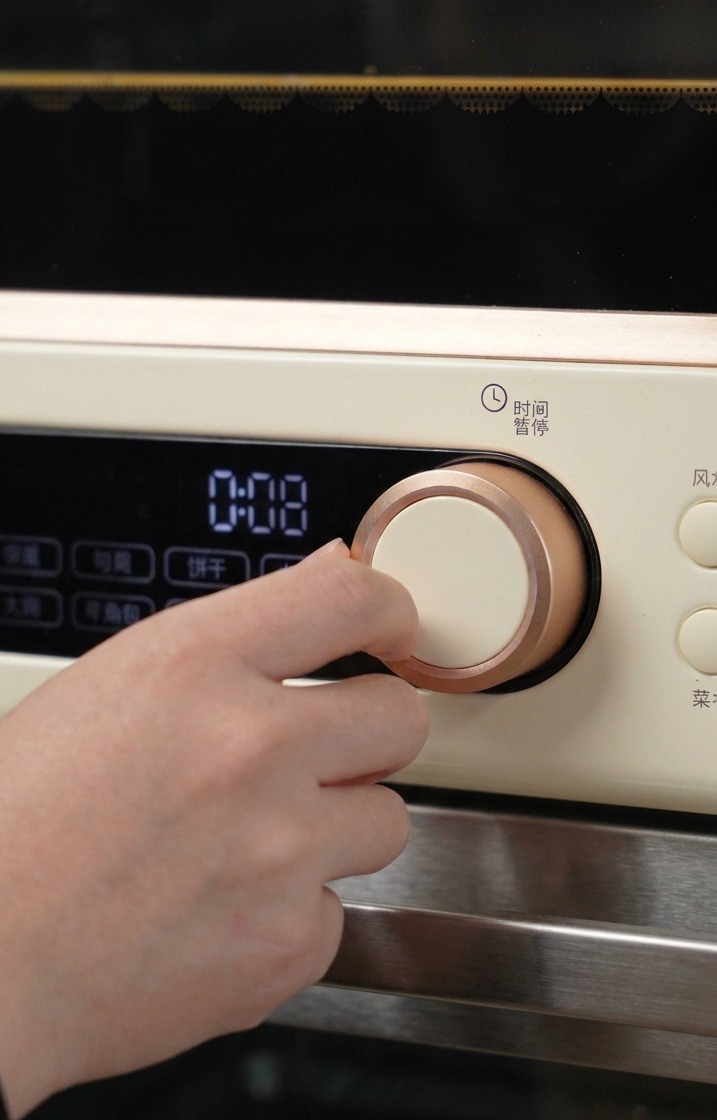 A hand turning the rose-gold dial of a cream-colored oven to set the initial baking time and temperature.
