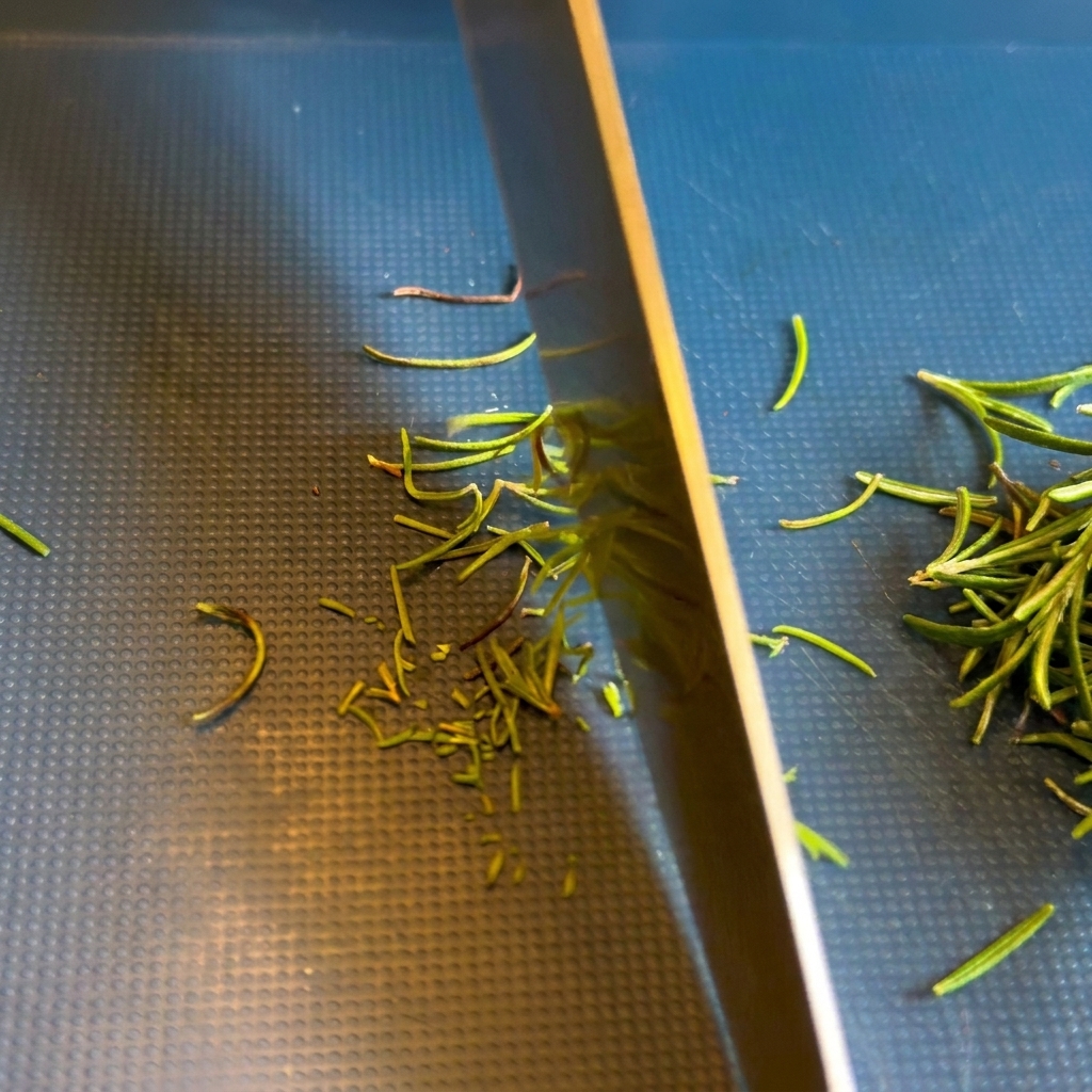 A close-up shot of a chefs knife blade mincing green rosemary needles on a dark, textured cutting board.