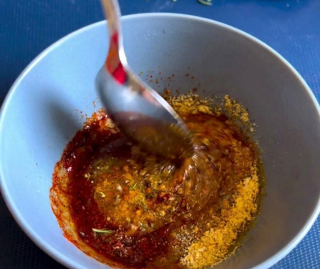 A metal spoon stirring a thick, vibrant red-brown spice and herb paste inside a small blue ceramic bowl.
