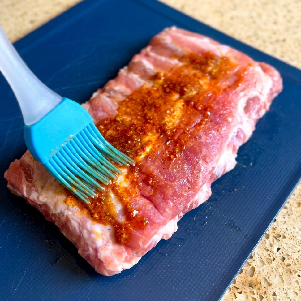 A blue silicone brush spreading a thick, grainy red marinade across the surface of a raw rack of pork ribs on a blue board.