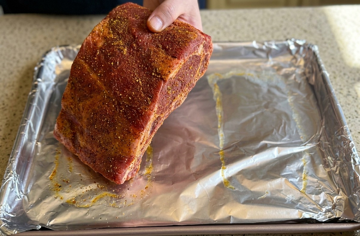 A hand placing a large, seasoned rack of ribs onto a baking sheet covered in crinkled silver aluminum foil.
