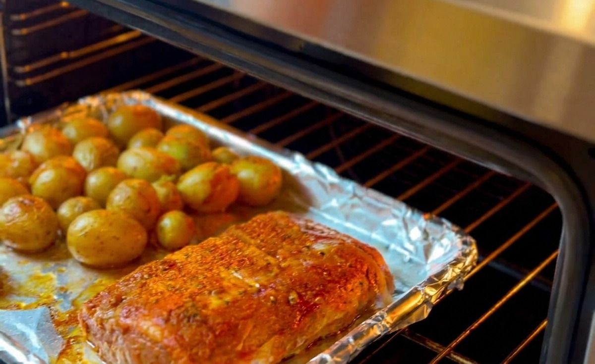 A baking tray with herb-rubbed ribs and small yellow potatoes being slid into a preheated oven for roasting.
