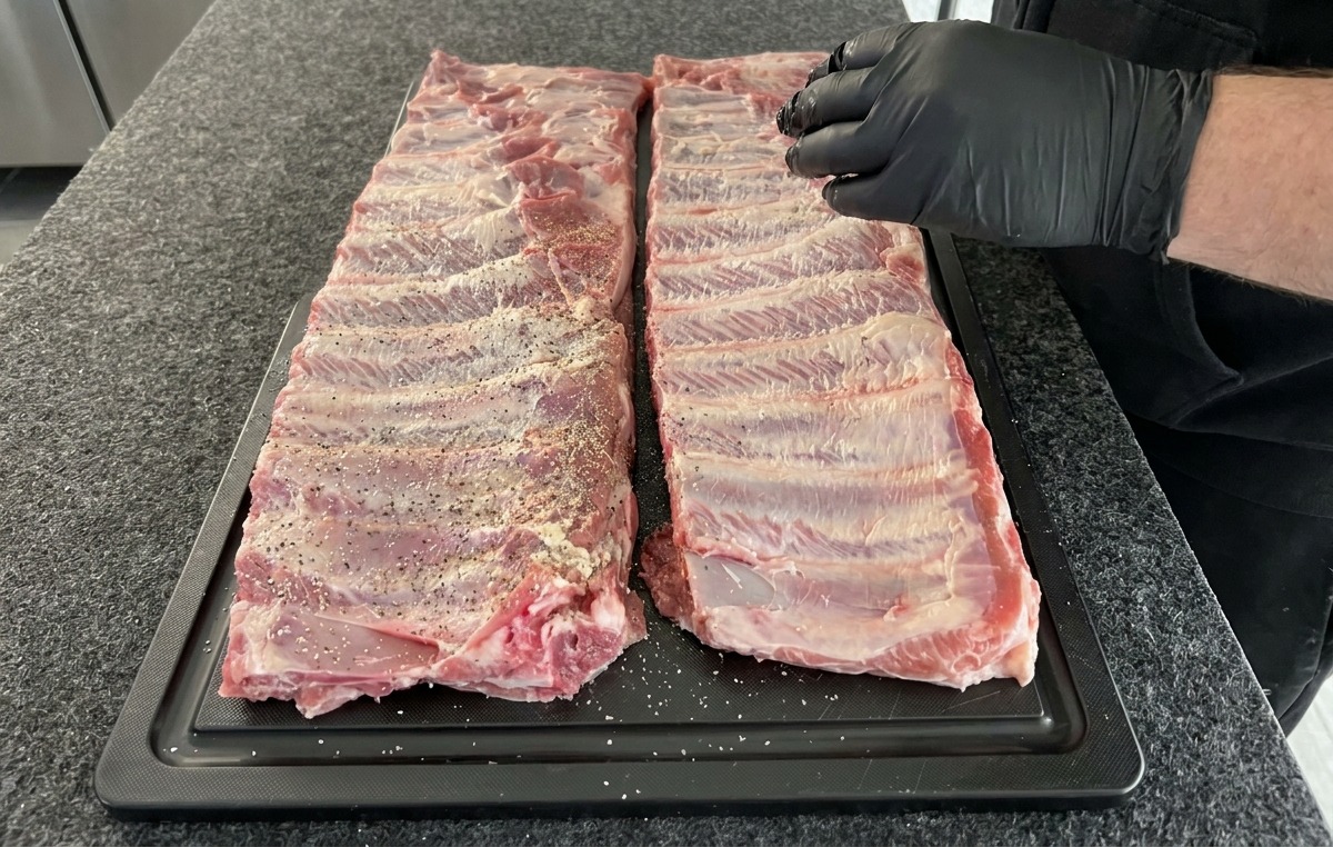 Two racks of raw pork ribs being seasoned with a dry rub of salt pepper and garlic powder on a black prep board.