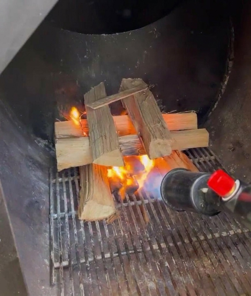 A propane torch being used to light a stack of split oak logs inside the metal firebox of a smoker.
