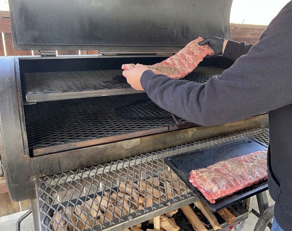 A person in a black hoodie and black gloves placing a rack of seasoned pork ribs onto the upper shelf of a large offset smoker.