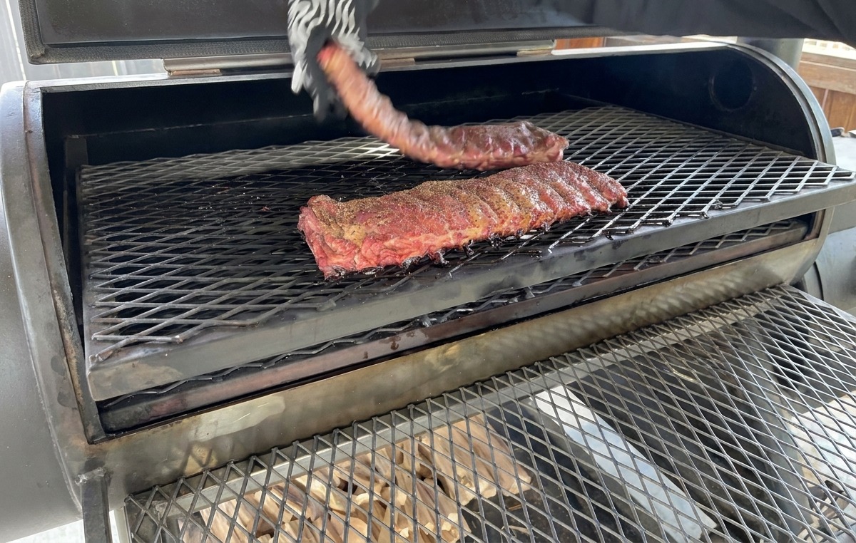 A hand wearing a patterned heat-resistant glove flipping a rack of partially smoked ribs on the metal grate of a smoker.