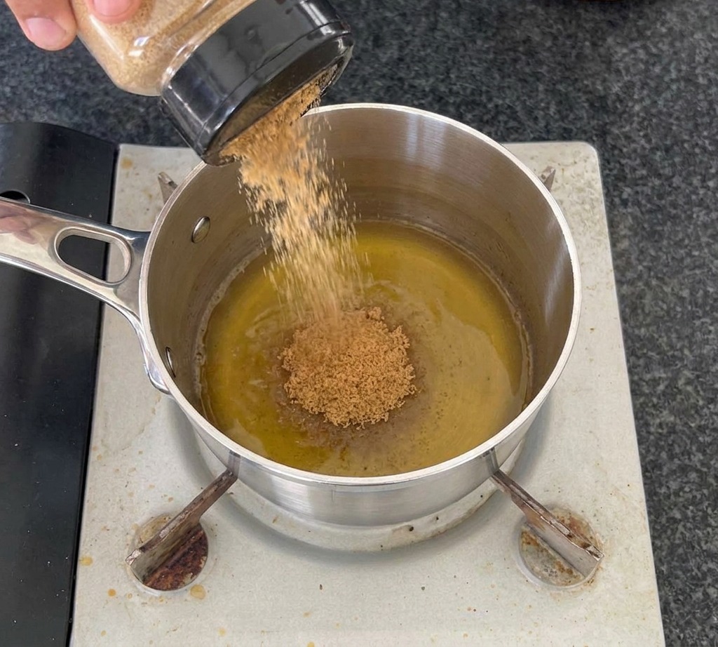 A hand pouring brown sugar from a clear jar into a stainless steel saucepan containing yellow apple cider vinegar on a portable burner.