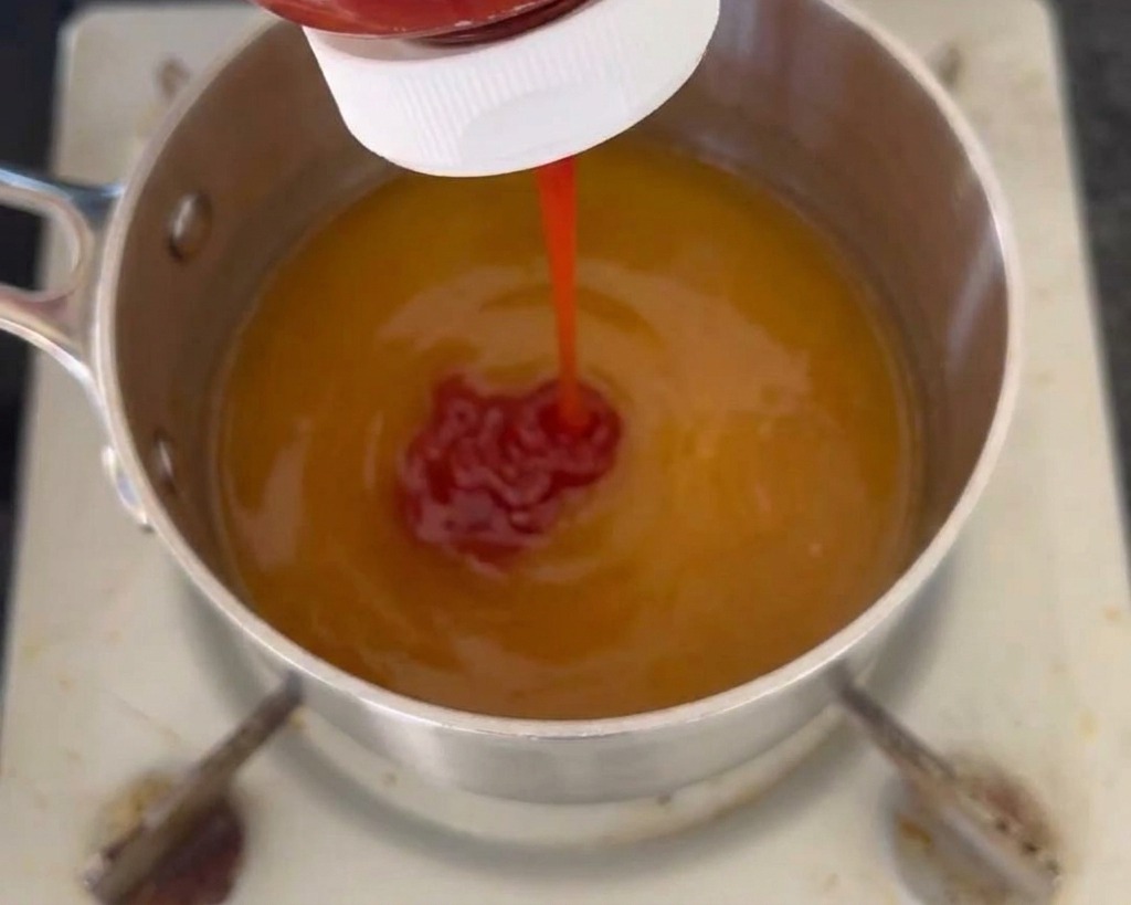A stream of red ketchup being squeezed from a bottle into a pot containing a yellow liquid sauce base.