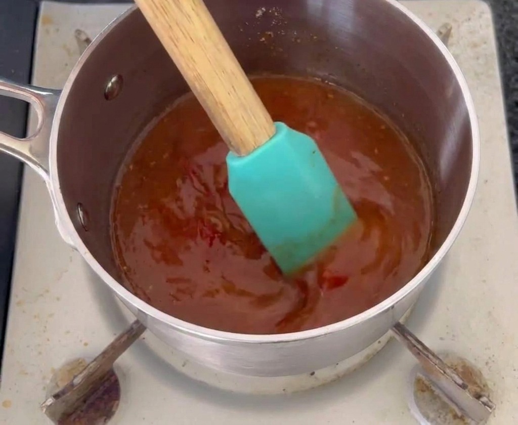 A teal silicone spatula stirring a dark orange-red mop sauce in a small stainless steel saucepan.