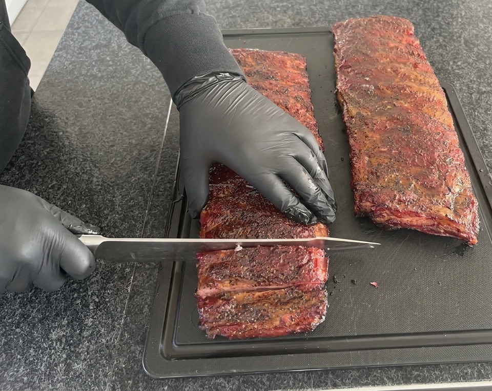 A person wearing black nitrile gloves using a large chef knife to slice through a long dark red smoked rack of ribs on a black cutting board.