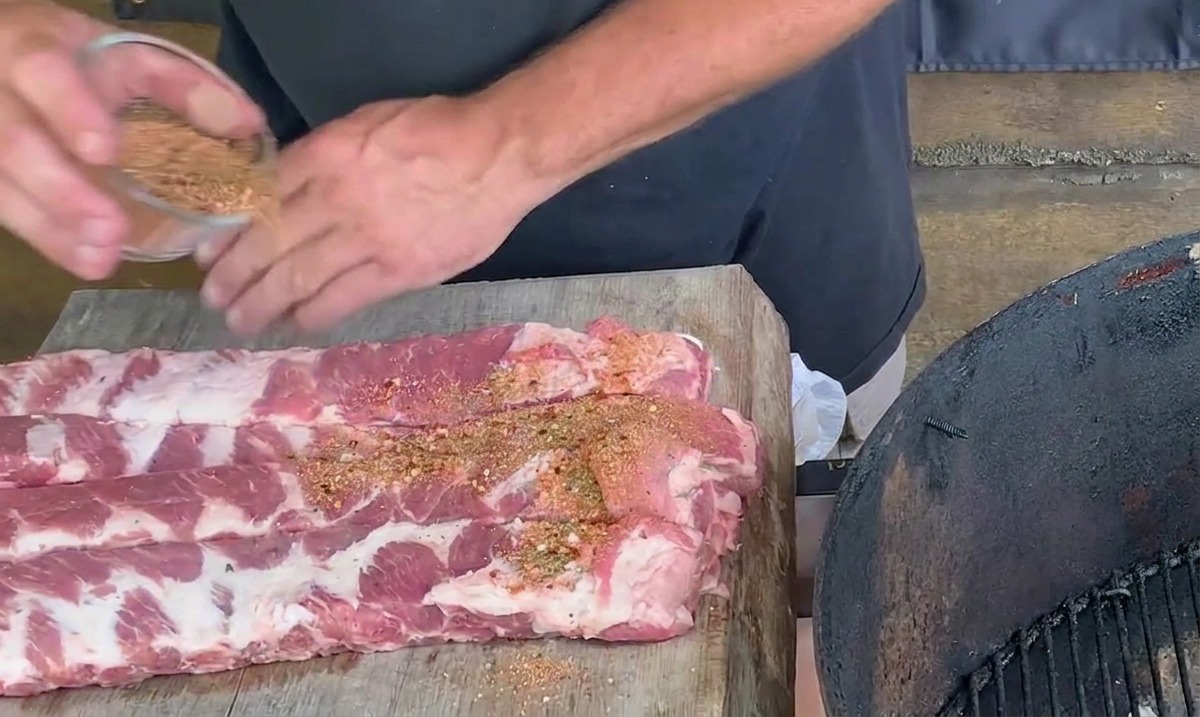 A persons hands sprinkling a reddish-brown spice mixture over raw pork ribs on a wooden cutting board.