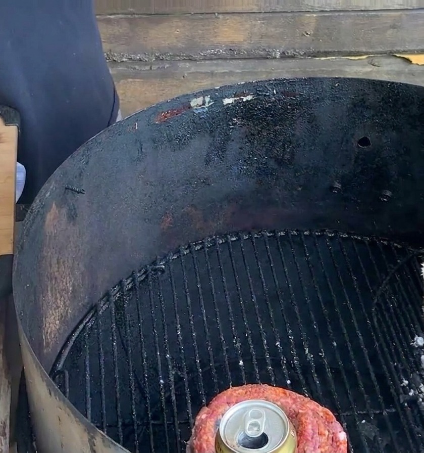 A tied rib cup containing a metal beer can sits on a black charcoal grill grate next to a wooden preparation board.