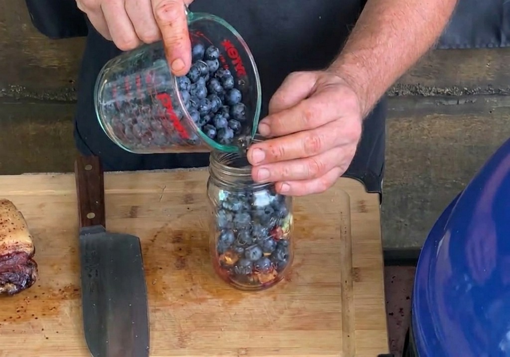 Fresh blueberries being poured from a glass measuring cup into a mason jar already containing chopped garlic and bacon bits.