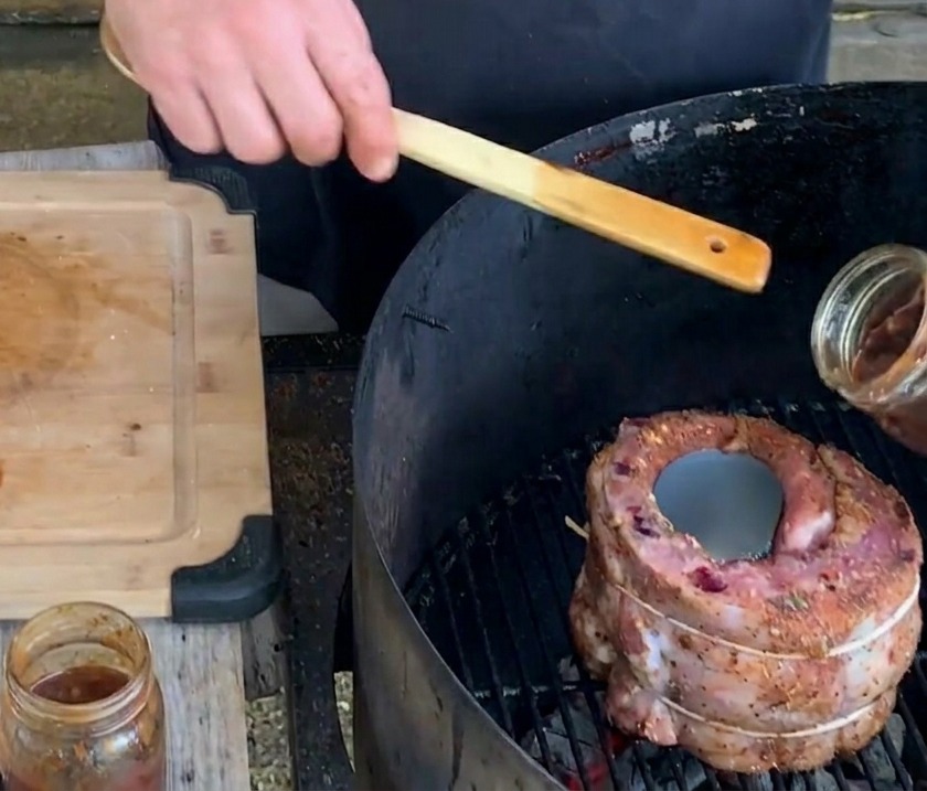 A hand pours a thick, dark blueberry sauce from a glass jar into a metal beer can centered inside a rib cup on the grill.