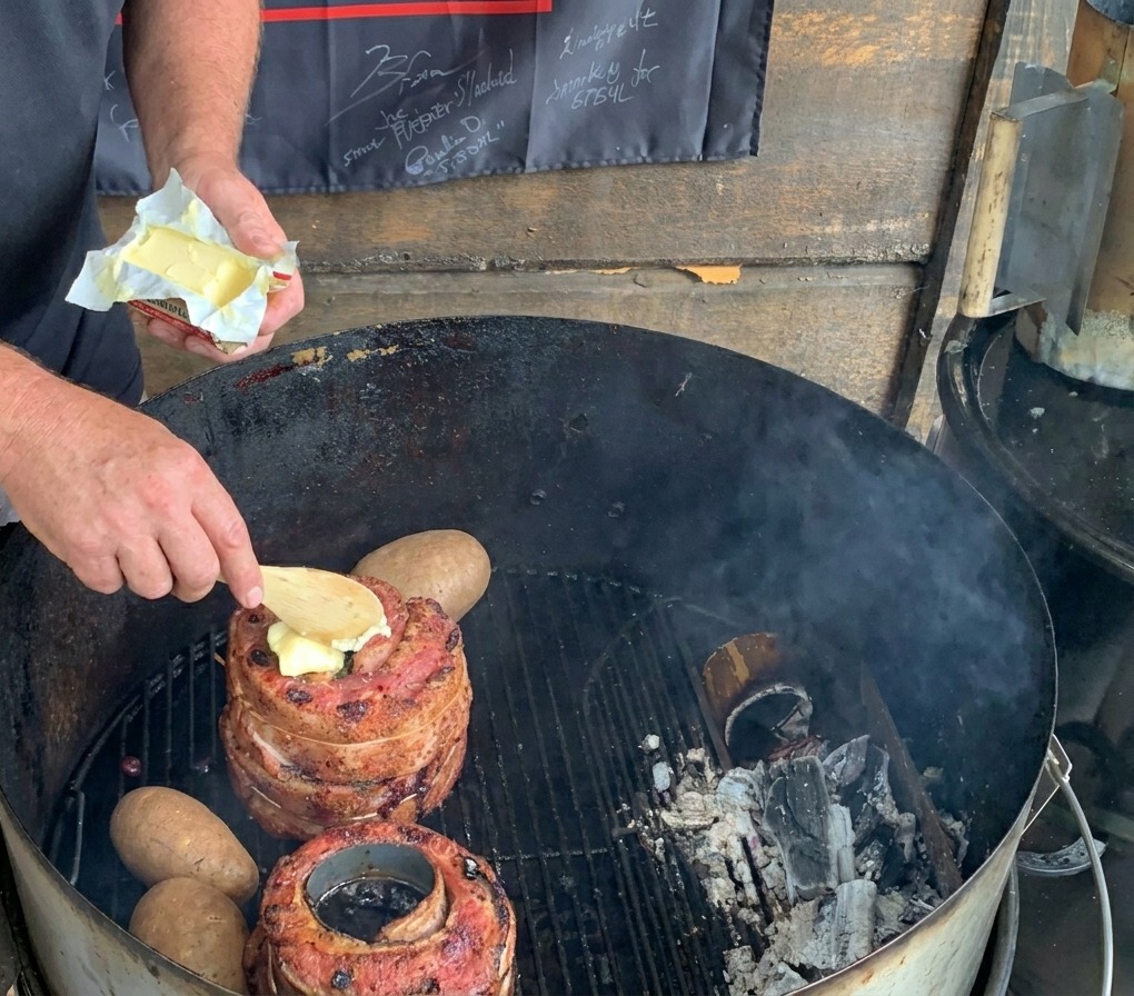 A wooden spoon placing a slice of butter onto meat on a charcoal grill next to whole potatoes.
