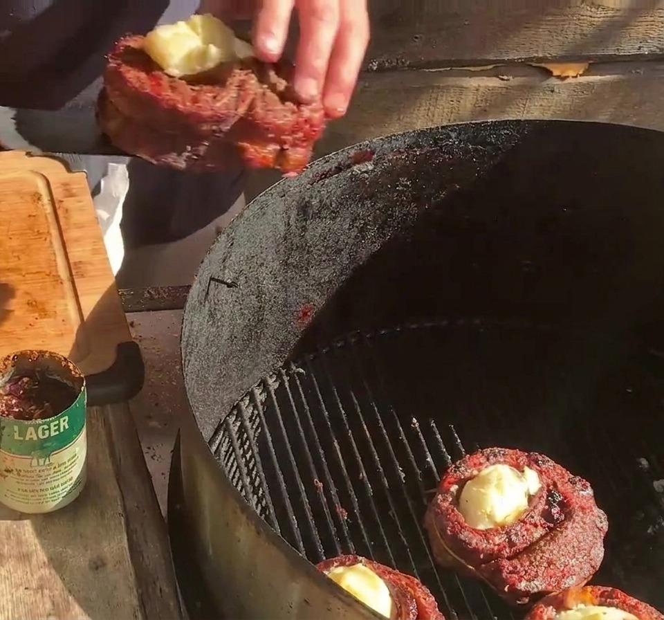 A person placing a stuffed rib cup onto a grill grate next to other rib cups and strips of bacon.