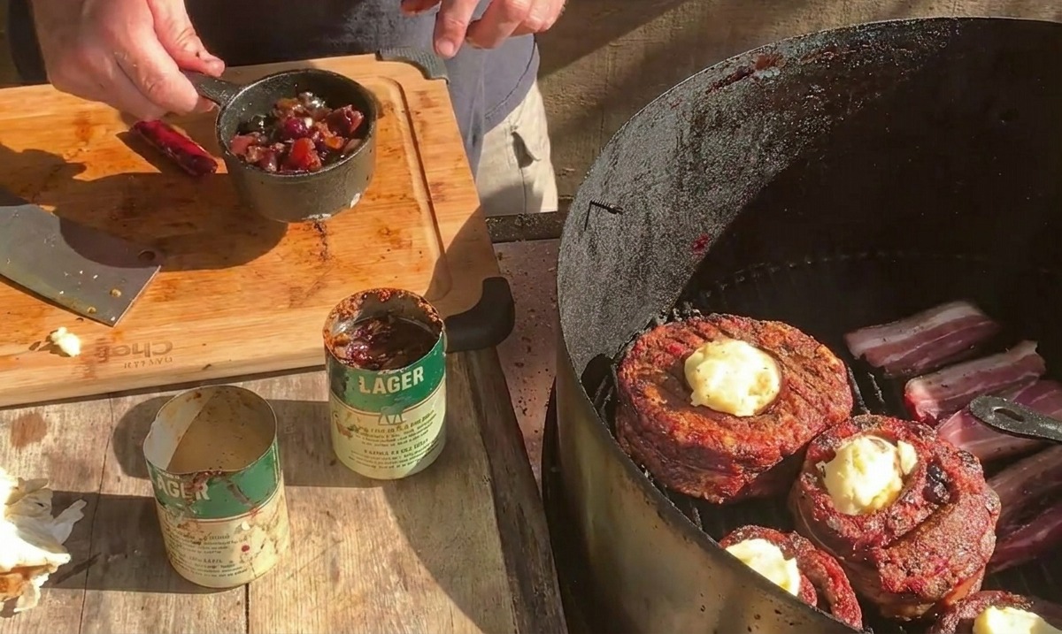 A person holding a small metal pot filled with blueberry sauce over a grill where bacon-wrapped rib cups are being topped with cheese.