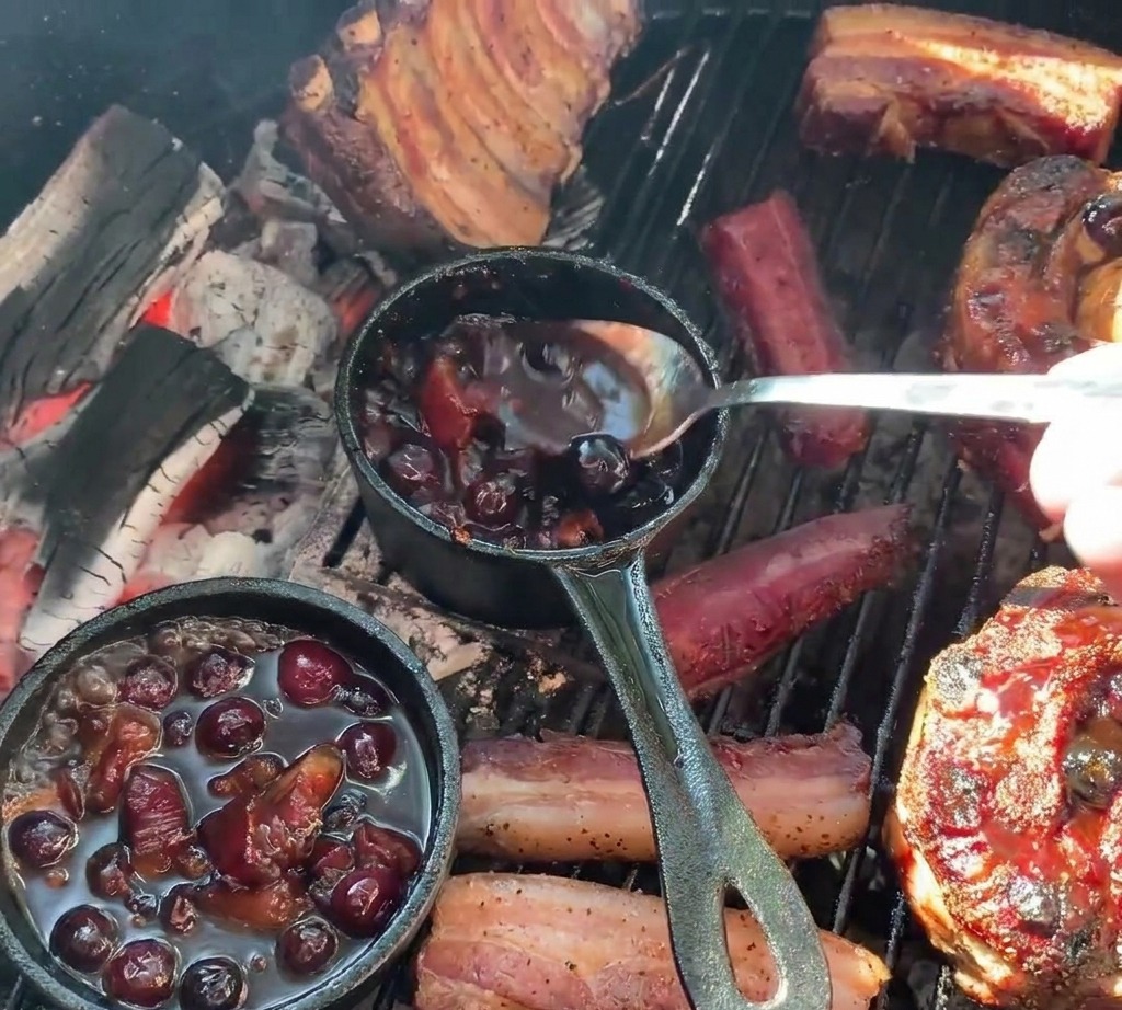 A person using a spoon to ladle dark blueberry sauce from a small cast iron pot onto grilled rib cups filled with potatoes.